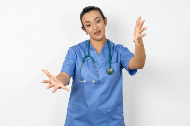 Arap doktor kadın mavi üniforma giyiyor kameraya bakıyor sarılmak için kollarını açmış gülümsüyor. Mutluluğu kucaklayan neşeli bir ifade..