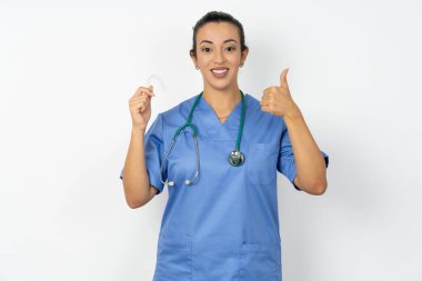 Üniformalı Arap kadın doktor, steteskop ile görünmez bir diş teli tutuyor ve baş parmağını kaldırıyor. Bu yeni tedaviyi öneriyor. Diş sağlığı konsepti.