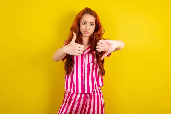 黄色いスタジオの背景にピンクのピンクのパジャマを身に着けている赤毛の女性は親指を上に示し 親指を下げ 難しい選択コンセプト — ストック写真