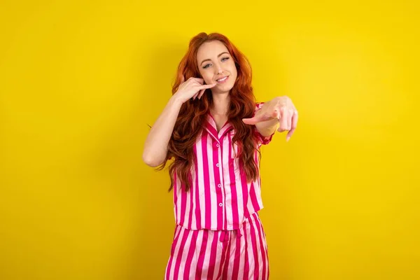 黄色いスタジオの背景にピンクのピンクのパジャマを着用した若い赤毛の女性は陽気で微笑み 後でジェスチャーを作りながらカメラを指差し 電話で話す — ストック写真