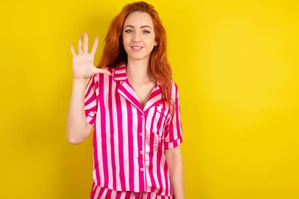 黄色いスタジオのバックグラウンドでピンクのピンクのパジャマを着た赤毛の女性が自信と幸せを笑いながら指の番号を5つ指で指で指摘します — ストック写真