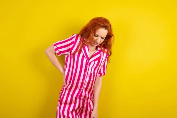 red haired woman wearing pink pyjama over yellow studio background suffering of backache, touching back with hand, muscular pain