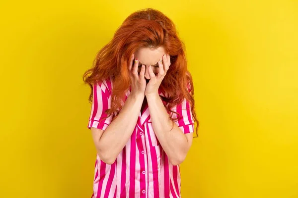 red haired woman wearing pink pyjama over yellow studio background covering her face with her hands, being devastated and crying. Sad concept