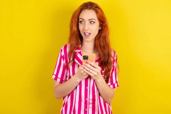 黄色いスタジオの背景にピンクのピンクのパジャマを身に着けている赤毛の女性は スマートフォンを保持し 空白のコピースペースで側面を見ています — ストック写真