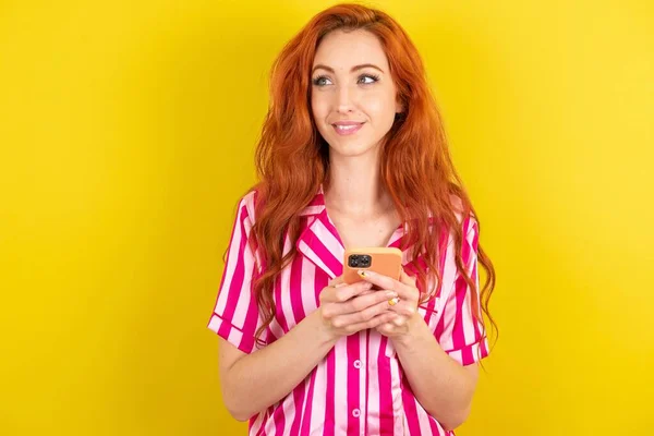 Rudowłosa Kobieta Różowej Piżamie Żółtym Tle Studio Trzymając Rękach Telefon — Zdjęcie stockowe