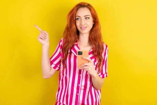 Улыбающаяся Рыжеволосая Женщина Розовом Пижаме Фоне Жёлтой Студии Держит Мобильный — стоковое фото