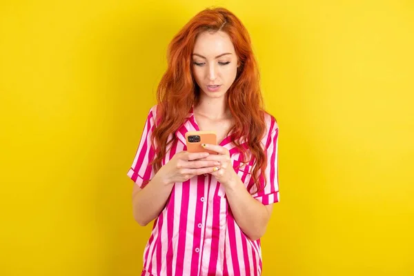 黄色いスタジオの背景にピンクのピンクのパジャマを着用した集中赤毛の女性は ソーシャルメディアニュースや重要な電子メールを読むスマートフォンを使用します — ストック写真