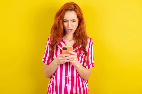 用智能手机阅读社交媒体新闻或重要的电子邮件 让人惊讶的红头发女人在黄色工作室的背景下穿着粉色睡衣 — 图库照片