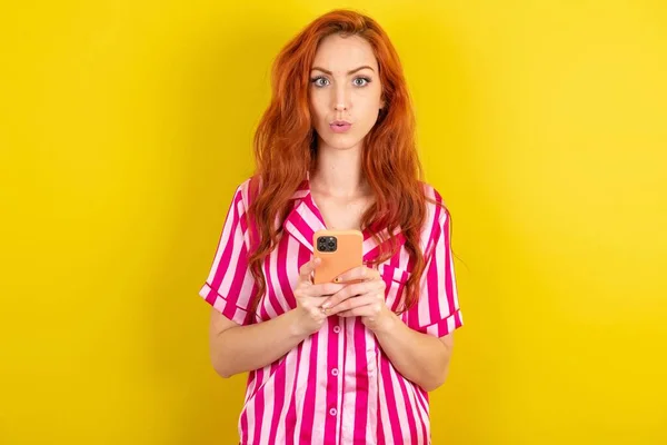 黄色いスタジオの背景にピンクのピンクのパジャマを身に着けているショックを受けた赤毛の女性は 信じられないほどの大きなショッピング価格を宣伝する電話読書を開きます — ストック写真