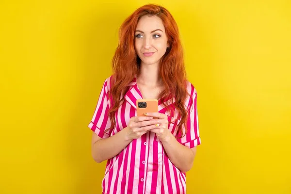 Rudowłosa Kobieta Różowej Piżamie Nad Żółtym Studio Tło Trzyma Telefon — Zdjęcie stockowe