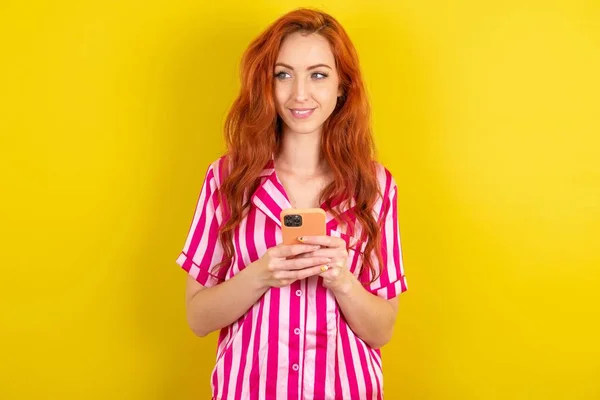 Рыжеволосая Женщина Розовом Пижаме Фоне Желтой Студии Держать Телефон Руки — стоковое фото