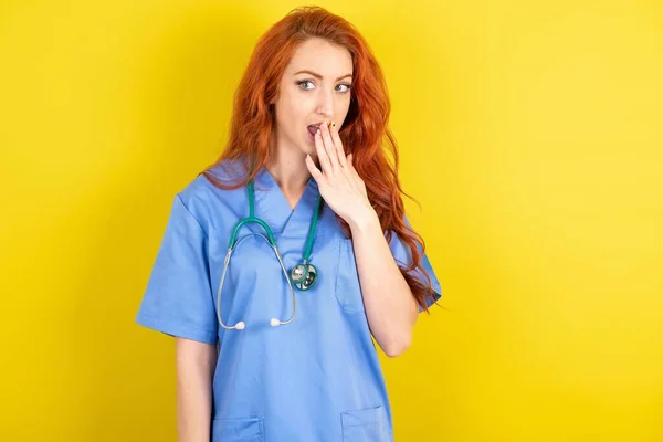 stock image young red-haired doctor woman covers mouth and looks with wonder at camera, cannot believe unexpected rumors.