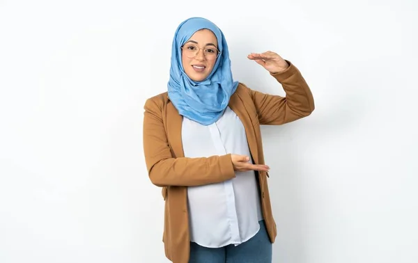 イスラム教徒の妊娠中の女性は 大きくて大きい看板を示す手でジェスチャーを着て シンボルを測定します カメラを見ている笑顔 — ストック写真