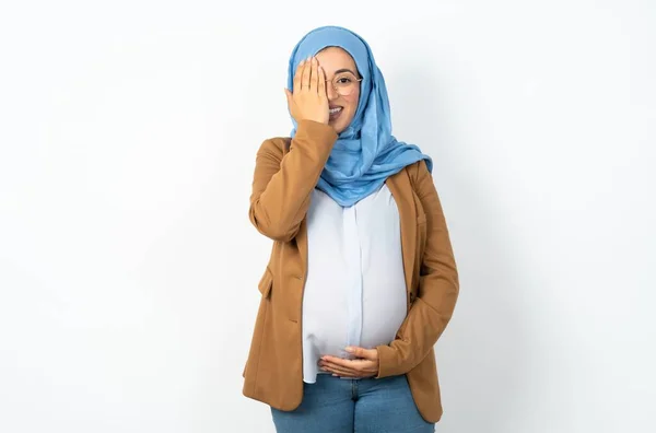 彼女の手で片目を覆うヒジャブを身に着けているイスラム教徒の妊婦 顔に自信を持って笑顔と驚きの感情 — ストック写真
