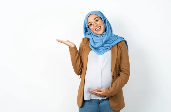 イスラム教徒の妊婦は 笑顔で陽気なプレゼンテーションを着て カメラを見ている手のひらを指す — ストック写真