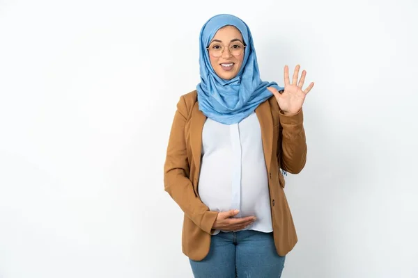 ヒジャーブを身に着けているイスラム教徒の妊婦は自信を持って幸せに微笑んでいる間指番号5を指で指で指さす — ストック写真