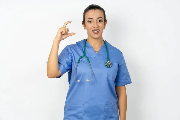 アラブの医師の女性は 青い制服を着て笑顔で自信を持って 指とカメラで小さなサイズのサインをする 測定コンセプト — ストック写真
