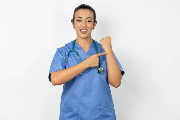 アラブの医師の女性は リラックスした表情でカメラを見ながら腕時計 忍耐を指して急いで青い制服を着ています — ストック写真