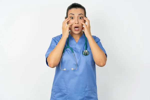 青い制服を着たスタイリッシュなアラブの医師の女性は興奮とスリルを表現し 顎を落とし 頬に手を当て 目が飛び出した — ストック写真