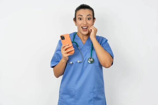 アラブの医師の女性は携帯電話装置が付いている青い制服のポーズを身に着け 現代スマートフォンのテキスト メッセージをタイプし 自由な時間の間に面白いビデオを見ます よいインターネット接続を楽しんで下さい — ストック写真