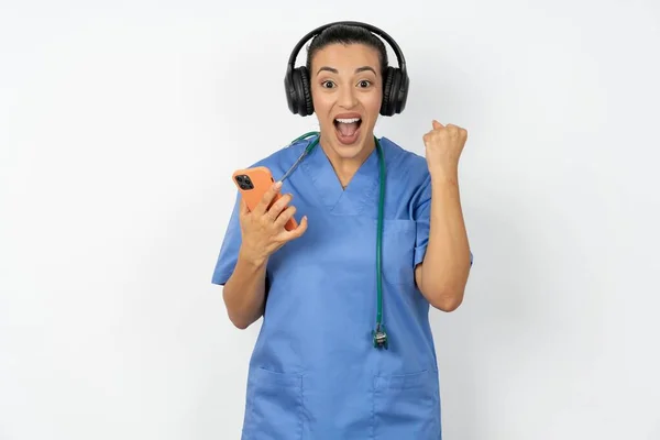 身穿蓝色制服的积极的阿拉伯医生女人拿着与耳机相连的现代手机 从美好的情感中握紧拳头 欢呼雀跃 — 图库照片
