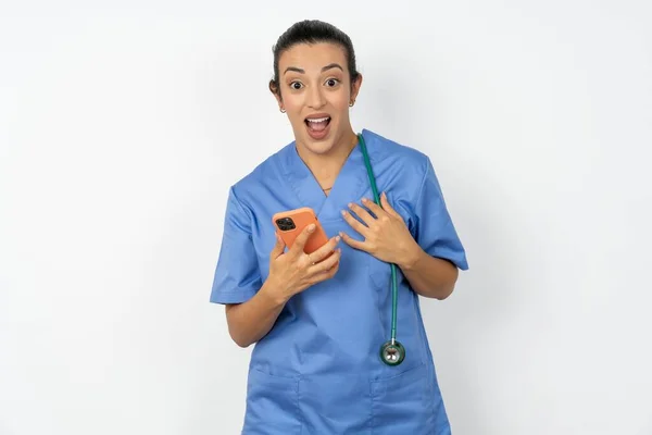 兴奋的阿拉伯医生女人身穿蓝色制服 手持智能手机 在收到好消息后 看着相机惊讶极了 — 图库照片