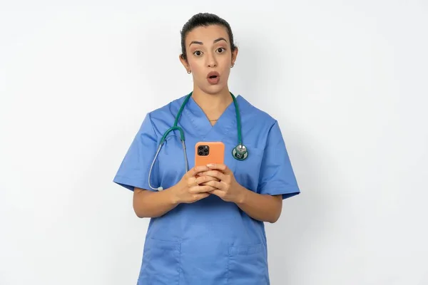 身穿蓝色制服的震惊的阿拉伯医生女人张开嘴拿着电话阅读广告难以置信的大购物价格 — 图库照片