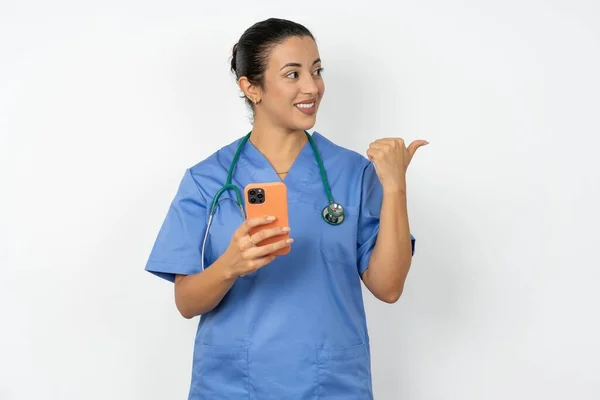 アラブの医師の女性は スマートフォンの指で青い制服を着てテキストし 幸せな顔の笑顔で親指で顔を示しています — ストック写真