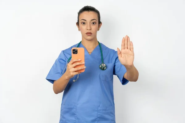 穿着蓝色制服的阿拉伯医生女士 使用智能手机并发短信 用张开的手做停止手势 表情严肃而自信 并做出防御姿态 — 图库照片