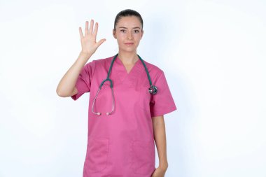 Pembe üniformalı beyaz kadın doktor. Steteskopla beş numarayı gösteriyor ve işaret ediyor. Kendinden emin ve mutlu bir şekilde gülümsüyor..