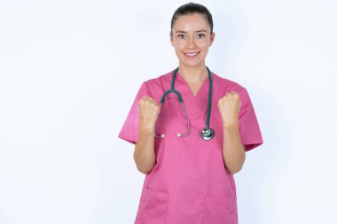 Pembe üniformalı neşeli beyaz kadın doktor steteskopla tezahürat yapıyor, başarıyı kutluyor, yumruklarını sıkarak evet diye bağırıyor.