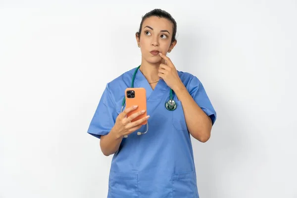 青い制服を着ているアラブの医師の女性は 何かについて深く考え 現代の携帯電話を使用して 良いメッセージを構成しようとし 唇の近くにインデックス指を保ちます — ストック写真