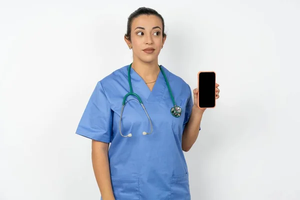 青い制服を着ているアラブの医師の女性は新しい携帯電話を保持し 現代の細胞の空白の表示を示す不思議な脇に見えます — ストック写真