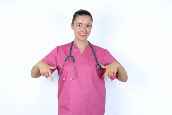 ポジティブな表情を持つピンクのユニフォームの白人女性医師は 両方の指で指を指し 幅広い興味のある笑顔を持っています そこを見てください — ストック写真