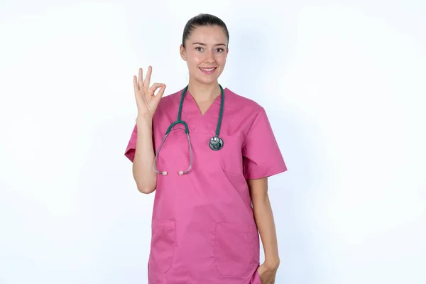 ステートスコープとピンクのユニフォームの白人女性医師は 表現承認として手でOkのサインを示し 楽観的である 陽気な表現を持っています — ストック写真