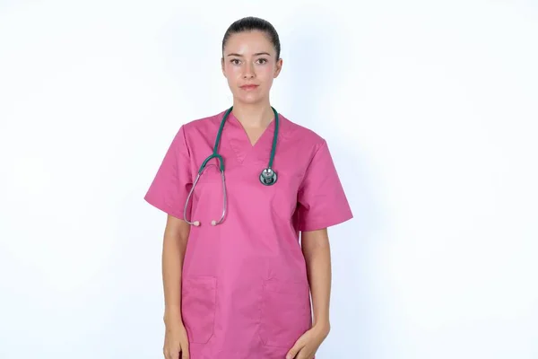 カメラを眺めながら 何かを考えるピンクのユニフォームの女性医師 両方の腕を下げ 中立的な表情 — ストック写真