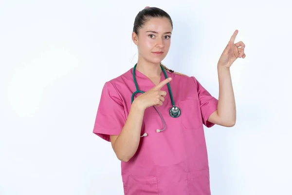ポジティブな表情を持つピンクのユニフォームの白人女性医師は あなたのプロモーションテキストや広告のための空白のコピースペースで指で示しています — ストック写真