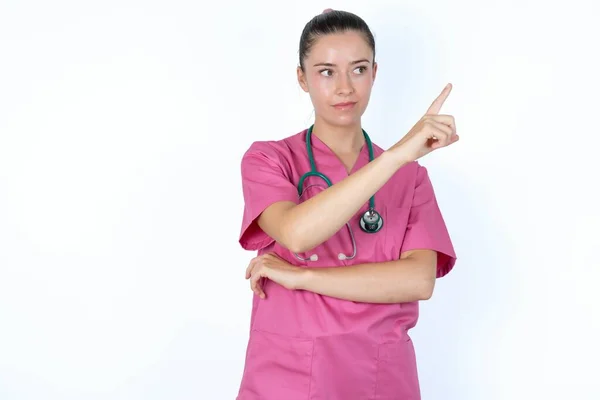 穿着粉色制服的高加索女医生的画像 在相机上摆出棘手的样子 用食指展示产品 广告概念 — 图库照片