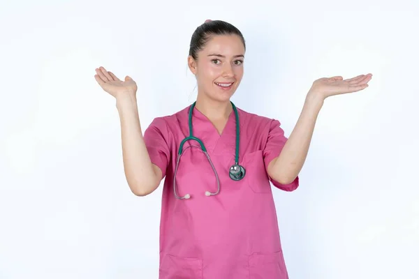 ステスコープとピンクのユニフォームで陽気な慎重な女性の医師は 頭の上に腕を上げる歓迎ジェスチャーを作ります — ストック写真