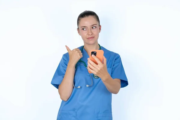 白人女性の医師は親指を指し 空白のスペースを脇に示し テキストメッセージを送信するための携帯電話を保持します — ストック写真