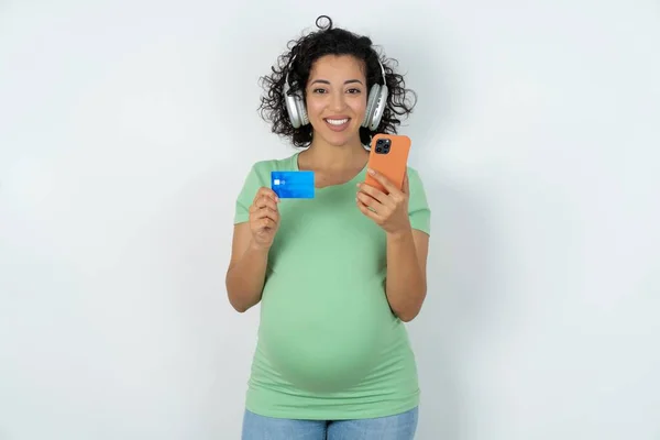 妊娠中の女性は耳にステレオヘッドフォンを着用し 現代の携帯電話とクレジットカードを保持しています — ストック写真