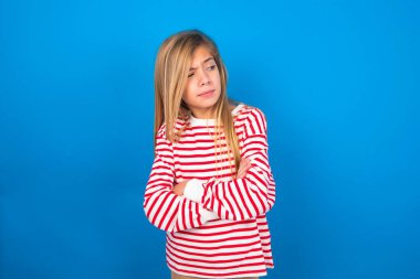 Mavi stüdyo geçmişine çizgili tişört giyen, kötü tavırlı, kollarını yana doğru kavuşturmuş güzel beyaz bir kız. Negatif insani duygu yüz ifadesi.