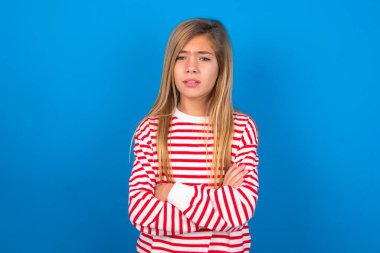 Güzel beyaz kız, mavi stüdyo arka planında çizgili tişört giyiyor, suratını hoşnutsuzca asıyor, kollarını kavuşturup bir açıklama bekliyor..