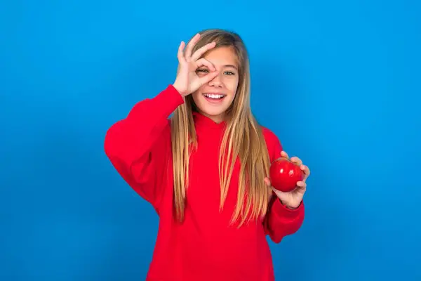 美しい白人十代の少女は 青い壁の上に赤いセーターを着用し 幸せな顔笑顔で指を通して見て手でOkのサインを行います — ストック写真