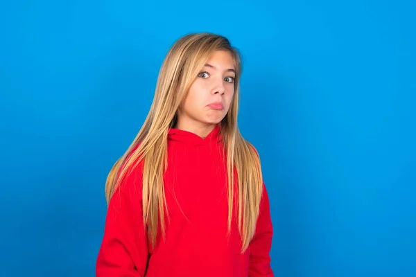美しい白人の十代の少女は 唇を曲げ 疑わしく懐疑的な表現 疑いと疑念を持って見て 眉を上げて青い壁の上に赤いセーターを着用しています — ストック写真