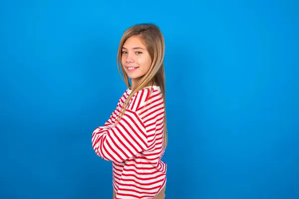 腕を交差した青いスタジオの背景にストライプのシャツを着ている陽気な美しい白人十代の少女のイメージ カメラを見たり笑ったり 信頼のコンセプト — ストック写真