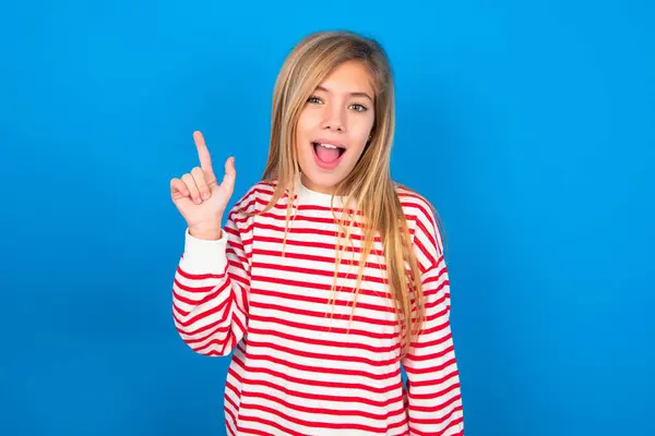 青いスタジオの背景にストライプのシャツを着ている美しい白人十代の少女が賢い表情を持ち 1本の指を上げ 重要なことを伝えることを忘れないように自分自身を覚えています — ストック写真