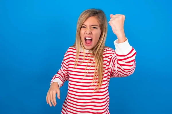 Fierce beautiful caucasian teen girl wearing striped shirt over blue studio background holding fist in front as if is ready for fight or challenge, screaming and having aggressive expression on face.