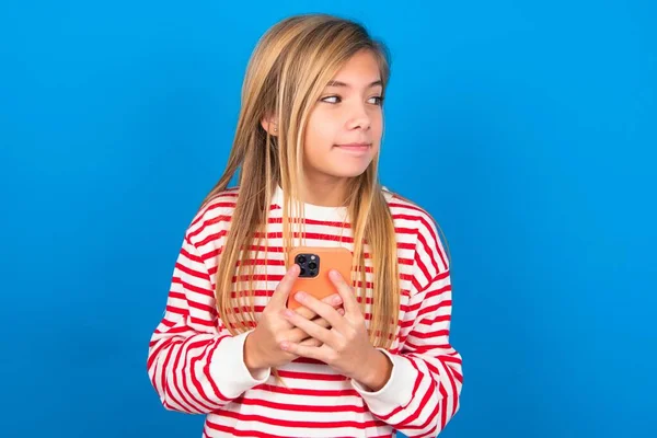 美しい慎重な十代の少女の肖像画は 夢のような外観で青いスタジオの背景にストライプのシャツを着て スマートフォンを保持しながら考えています メッセージを書くために試みる — ストック写真