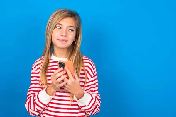 幸せな美しい白人少女は 携帯電話を使用してイヤホンで音楽を聞く青いスタジオの背景の上にストライプシャツを着用 — ストック写真